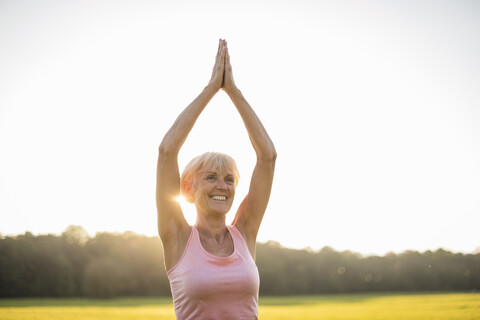 Lächelnde ältere Frau beim Yoga auf einer ländlichen Wiese bei Sonnenuntergang, lizenzfreies Stockfoto