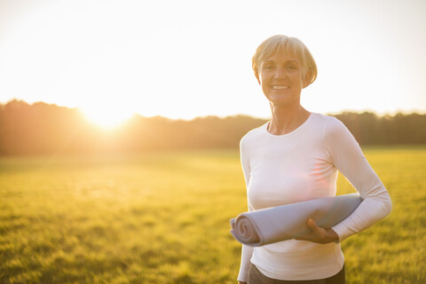 Porträt einer lächelnden älteren Frau mit Yogamatte auf einer ländlichen Wiese bei Sonnenuntergang, lizenzfreies Stockfoto