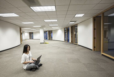 An Asian businesswoman sitting cross legged and working on a laptop computer in the middle of an empty office space. - MINF09545