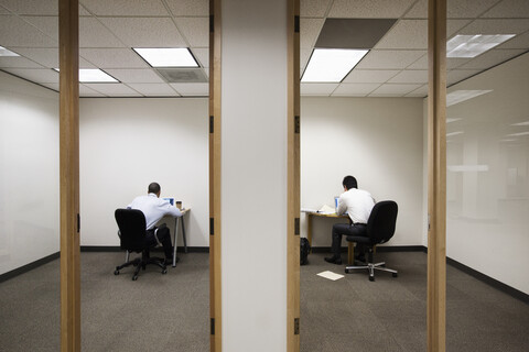 Blick auf zwei Geschäftsleute, die auf gegenüberliegenden Seiten einer Wand in zwei verschiedenen Büroräumen arbeiten., lizenzfreies Stockfoto