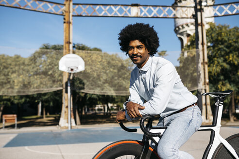 Lächelnder Mann, der auf seinem Fahrrad auf einem Basketballplatz sitzt - JRFF01965