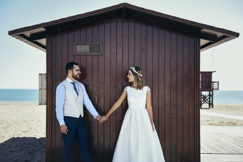 Brautpaar steht Hand in Hand vor einer Strandhütte und schaut sich an, lizenzfreies Stockfoto