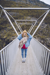Junge Frau steht auf einer windigen Brücke und hält ihre Mütze - RSGF00090