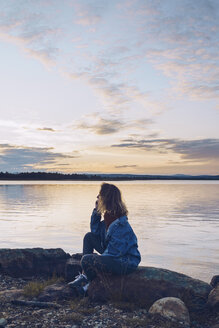 Junge Frau sitzt am Inari-See und betrachtet die Aussicht, Finnland - RSGF00094