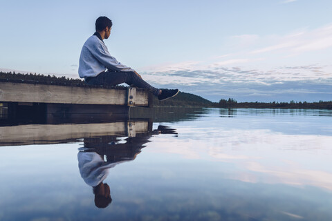 Junger Mann sitzt am Inari-See und schaut auf die Aussicht, Finnland, lizenzfreies Stockfoto