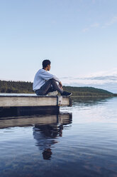 Junger Mann sitzt am Inari-See und schaut auf die Aussicht, Finnland - RSGF00096