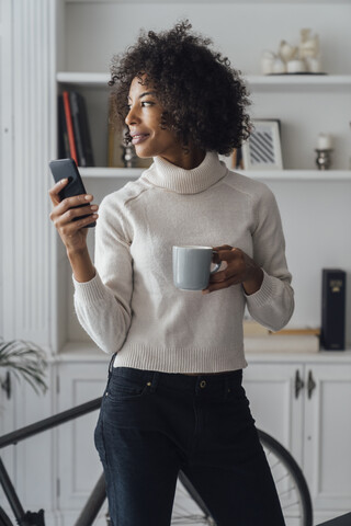 Mid adult freelancer standing in her home office, using smartphone, holding coffee cup stock photo