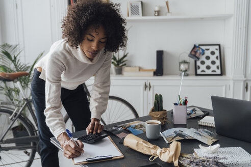 Freelancer standing at hert desk, using calculater, taking notes - BOYF00930