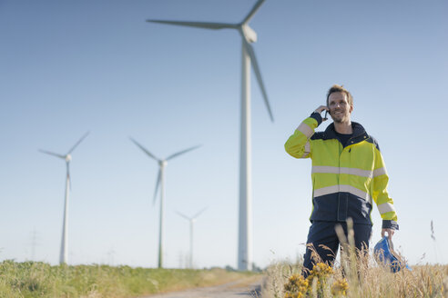 Lächelnder Ingenieur, der auf einem Feld in einem Windpark steht und mit einem Handy spricht - GUSF01305