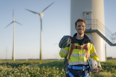 Porträt eines lächelnden Technikers, der in einem Windpark mit Kletterausrüstung steht - GUSF01342