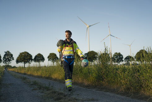 Technician walking on field path at a wind farm with climbing equipment - GUSF01345