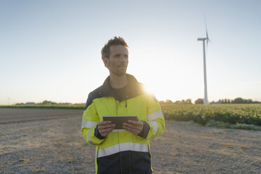 Ingenieur steht in ländlicher Landschaft an einer Windkraftanlage und hält eine Tafel - GUSF01348