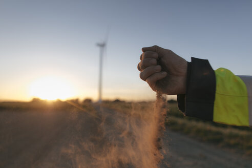 Nahaufnahme der Hand eines Mannes, die bei Sonnenuntergang Erde auf eine Windkraftanlage streut - GUSF01380