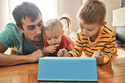 Vater und seine Kinder liegen zu Hause auf dem Boden und benutzen ein digitales Tablet, lizenzfreies Stockfoto