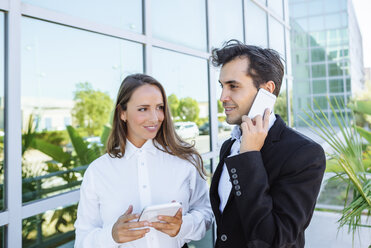 Lächelnde Geschäftsfrau und Geschäftsmann mit Tablet und Mobiltelefon vor einem Bürogebäude - KIJF02096