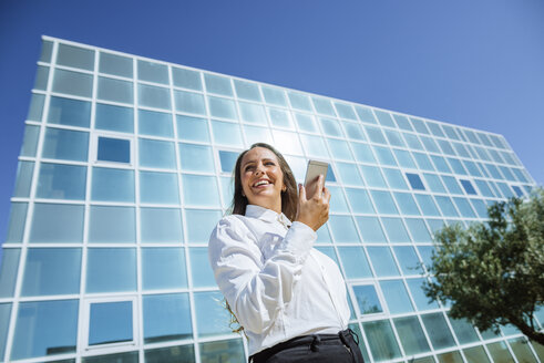 Lächelnde Geschäftsfrau, die ein Mobiltelefon vor einem Bürogebäude benutzt - KIJF02100