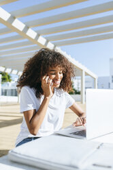 Lächelnde junge Frau am Telefon, die am Tisch sitzt und einen Laptop benutzt - KIJF02120