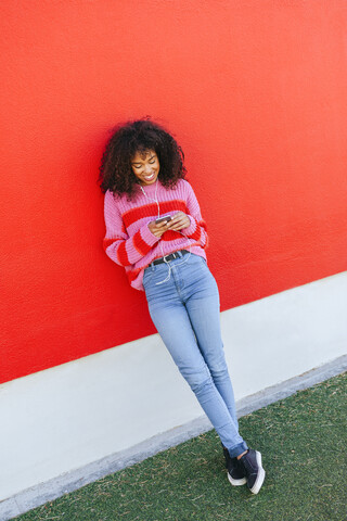 Smiling young woman with earphones leaning against red wall looking at cell phone stock photo