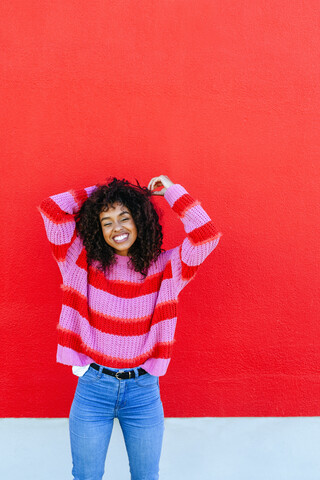 Porträt einer lachenden jungen Frau mit lockigem Haar vor einer roten Wand stehend, lizenzfreies Stockfoto
