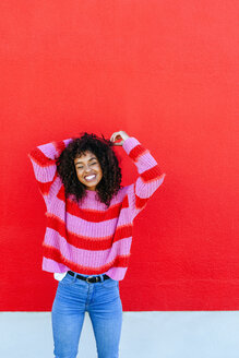 Portrait of laughing young woman with curly hair standing in front of red wall - KIJF02126