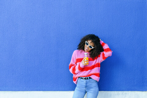 Porträt einer lächelnden jungen Frau mit einer Flasche Orangensaft vor einer blauen Wand stehend, lizenzfreies Stockfoto