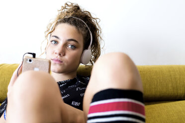 Porträt eines Mädchens, das mit Kopfhörern und Smartphone auf der Couch sitzt und Musik hört - ERRF00051