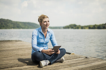 Frau sitzt auf einem Steg an einem See und benutzt ein Tablet - MOEF01485