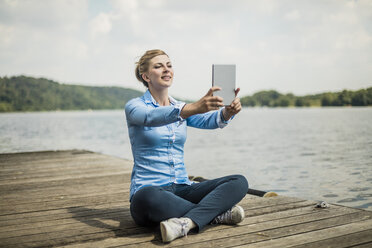 Lächelnde Frau sitzt auf einem Steg an einem See und macht ein Selfie mit einem Tablet - MOEF01486