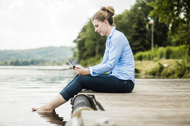 Frau sitzt auf einem Steg an einem See und benutzt ein Tablet - MOEF01492