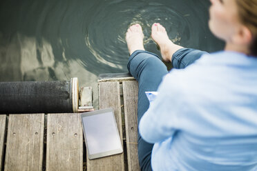 Frau sitzt auf Steg an einem See neben Tablet mit Füßen im Wasser - MOEF01496