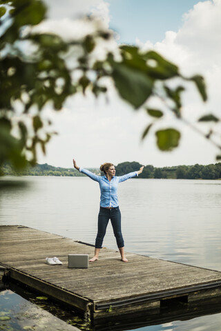 Frau streckt sich auf einem Steg an einem See neben einem Laptop, lizenzfreies Stockfoto