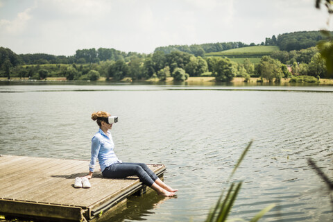 Woman with VR glasses sitting on jetty at a lake with feet in water stock photo