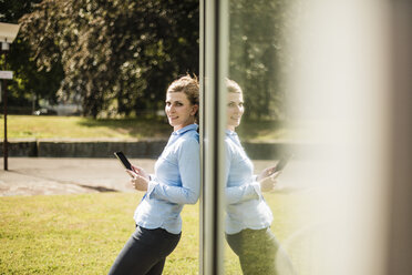 Smiling woman leaning against glass front holding tablet - MOEF01514