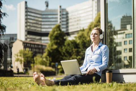 Frau mit Kopfhörern, die auf einer städtischen Wiese sitzt und einen Laptop benutzt, lizenzfreies Stockfoto