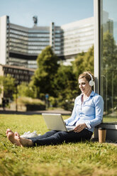 Smiling woman wearing headphones sitting on urban meadow using laptop - MOEF01518