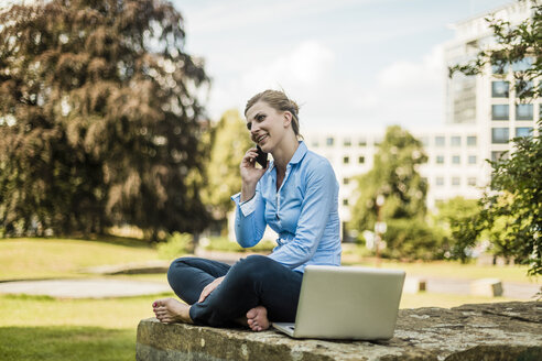 Lächelnde Frau, die in einem Stadtpark sitzt und mit einem Handy telefoniert - MOEF01519