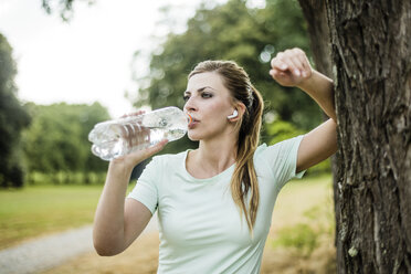 Sportliche junge Frau lehnt an einem Baum in einem Park und trinkt aus einer Flasche - MOEF01536