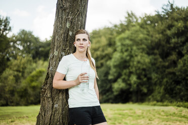 Sportliche junge Frau lehnt mit einer Flasche in der Hand an einem Baum in einem Park - MOEF01537