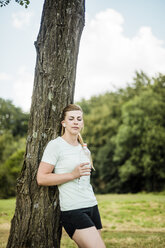 Sportliche junge Frau lehnt mit einer Flasche in der Hand an einem Baum in einem Park - MOEF01538