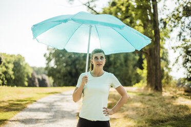 Sportliche junge Frau steht in einem Park und hält einen Sonnenschirm - MOEF01546