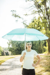 Sportliche junge Frau steht in einem Park und hält einen Sonnenschirm - MOEF01547
