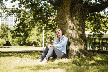 Frau sitzt in einem Stadtpark und lehnt sich gegen einen Baum und spricht mit einem Handy - MOEF01552