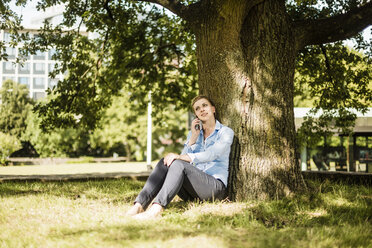 Woman sitting in urban park leaning against a tree talking on cell phone - MOEF01552