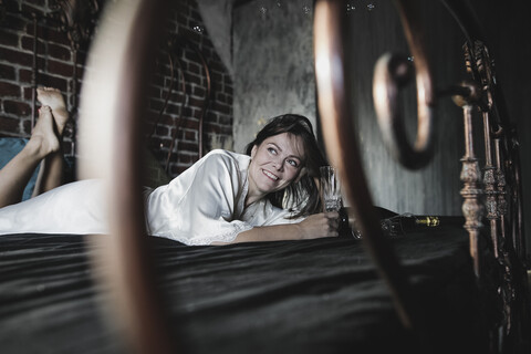 Lächelnde Frau auf dem Bett liegend mit einem Sektglas in der Hand, lizenzfreies Stockfoto