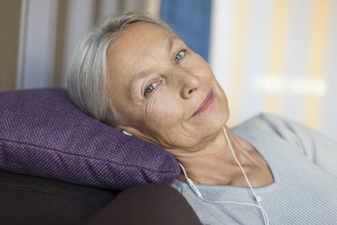 Porträt einer lächelnden älteren Frau, die zu Hause auf der Couch liegt und mit Kopfhörern Musik hört - VGF00121