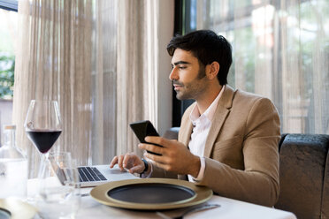 Man sitting at table in a restaurant using laptop and cell phone - VABF01645