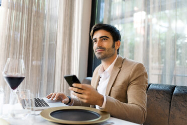 Smiling man sitting at table in a restaurant using laptop and cell phone - VABF01646