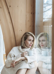 Frau mit Kaffeetasse schaut durch das Fenster, während sie zu Hause sitzt - CAVF54096