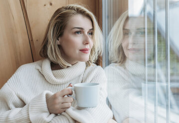 Woman holding coffee cup while looking through window at home - CAVF54097