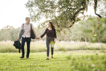 Lächelndes Paar hält sich an den Händen, während es auf einer Wiese im Park spazieren geht - CAVF54124